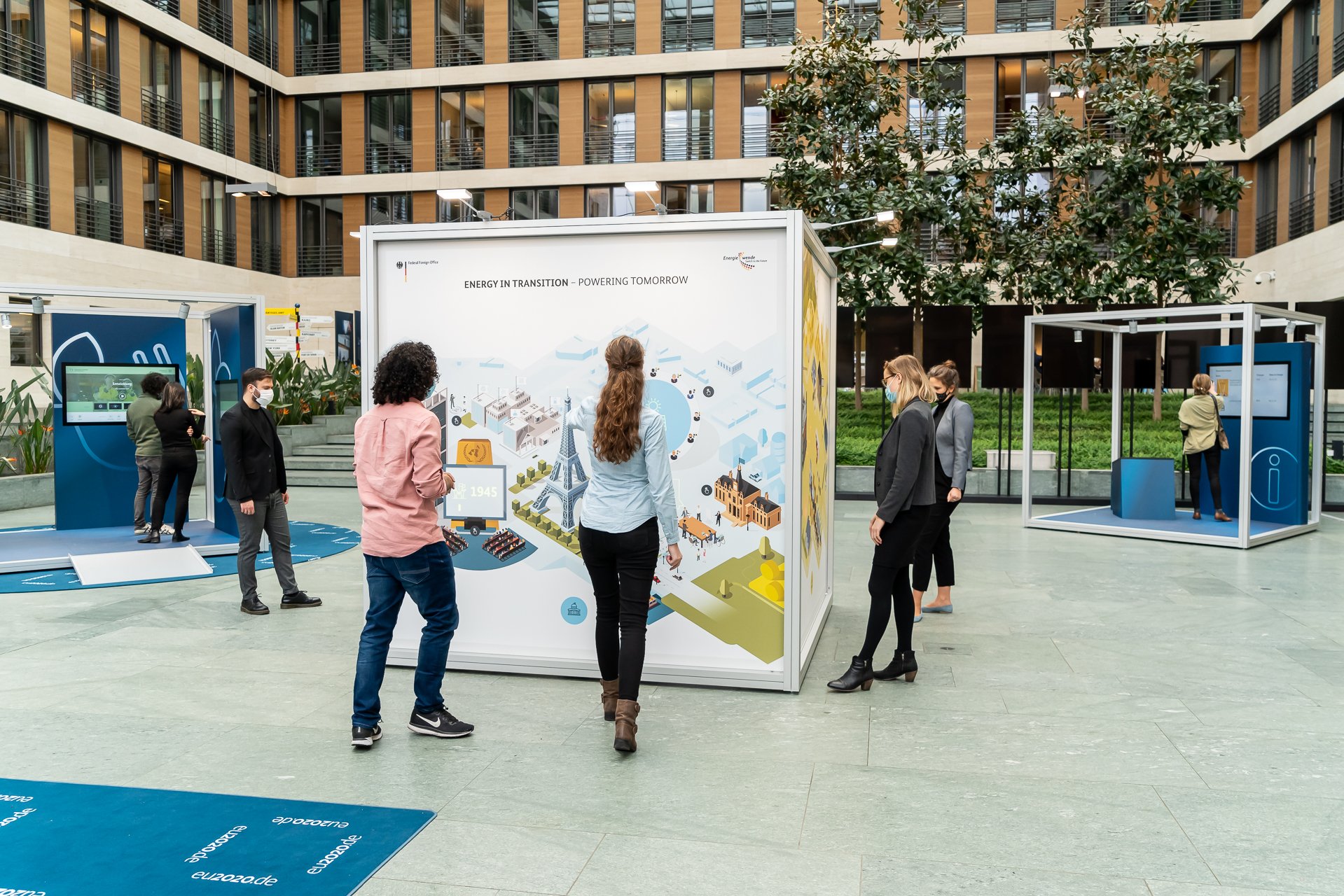 A snapshot of visitors at the travelling exhibition. The cube-shaped Energy in Transition station is in the foreground. Visitors are standing at the different sides of the cube and are pointing at and viewing the cube’s animations that are lighting up. In the background you can see visitors standing at the other stations.