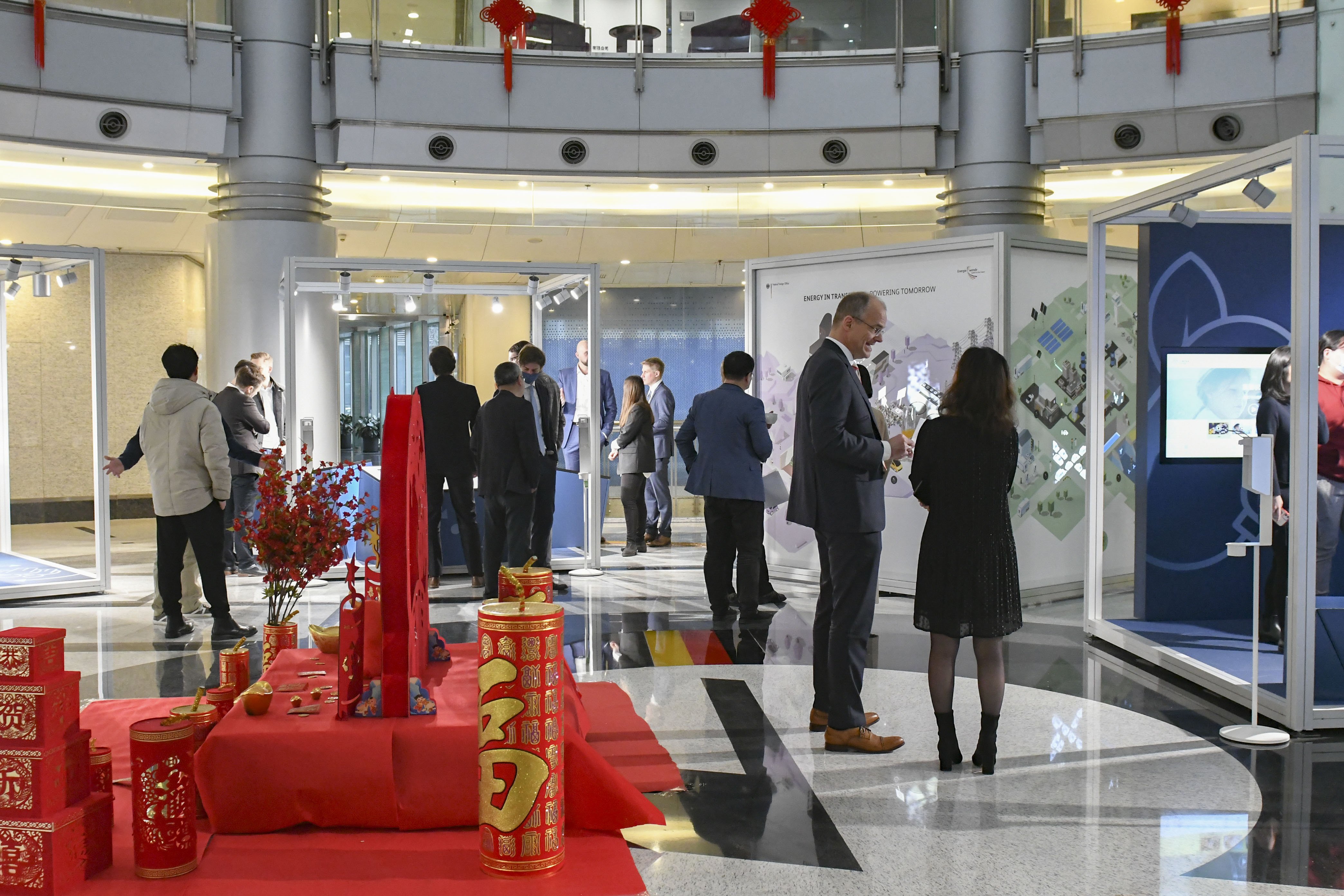 Besucher*innen stehen im Foyer des German Centers in Shanghai zur Eröffnung der Wanderausstellung. Sie schauen sich die Ausstellungselemente an. Zwei Männer unterhalten sich stehend neben roter chinesischer Dekoration. Januar 2021.