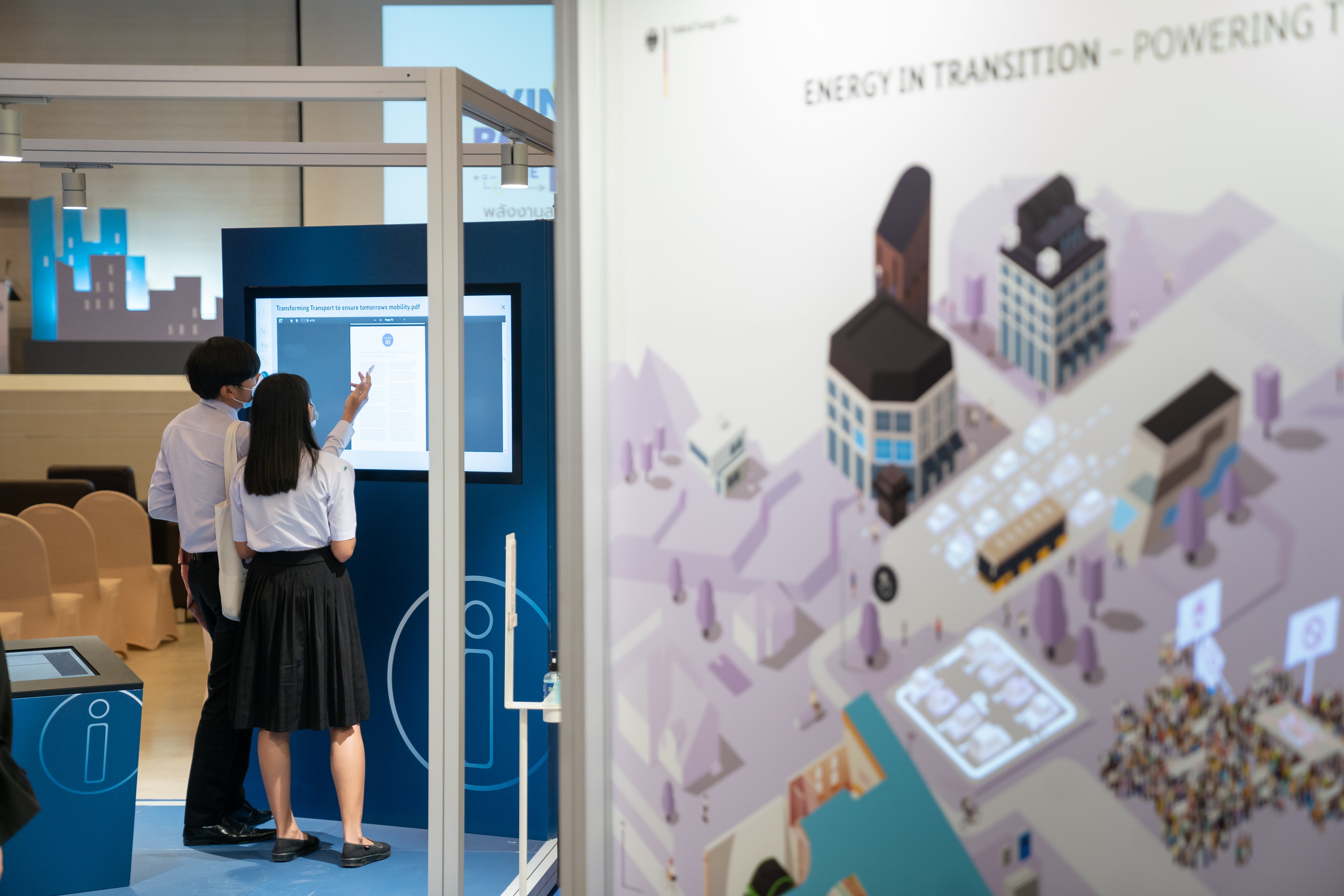In the forefront, the photo shows one side of the "Energy Transition Cube". In the background, there are a man and a woman standing at the "Info Lounge". Both are looking at the touch screen.