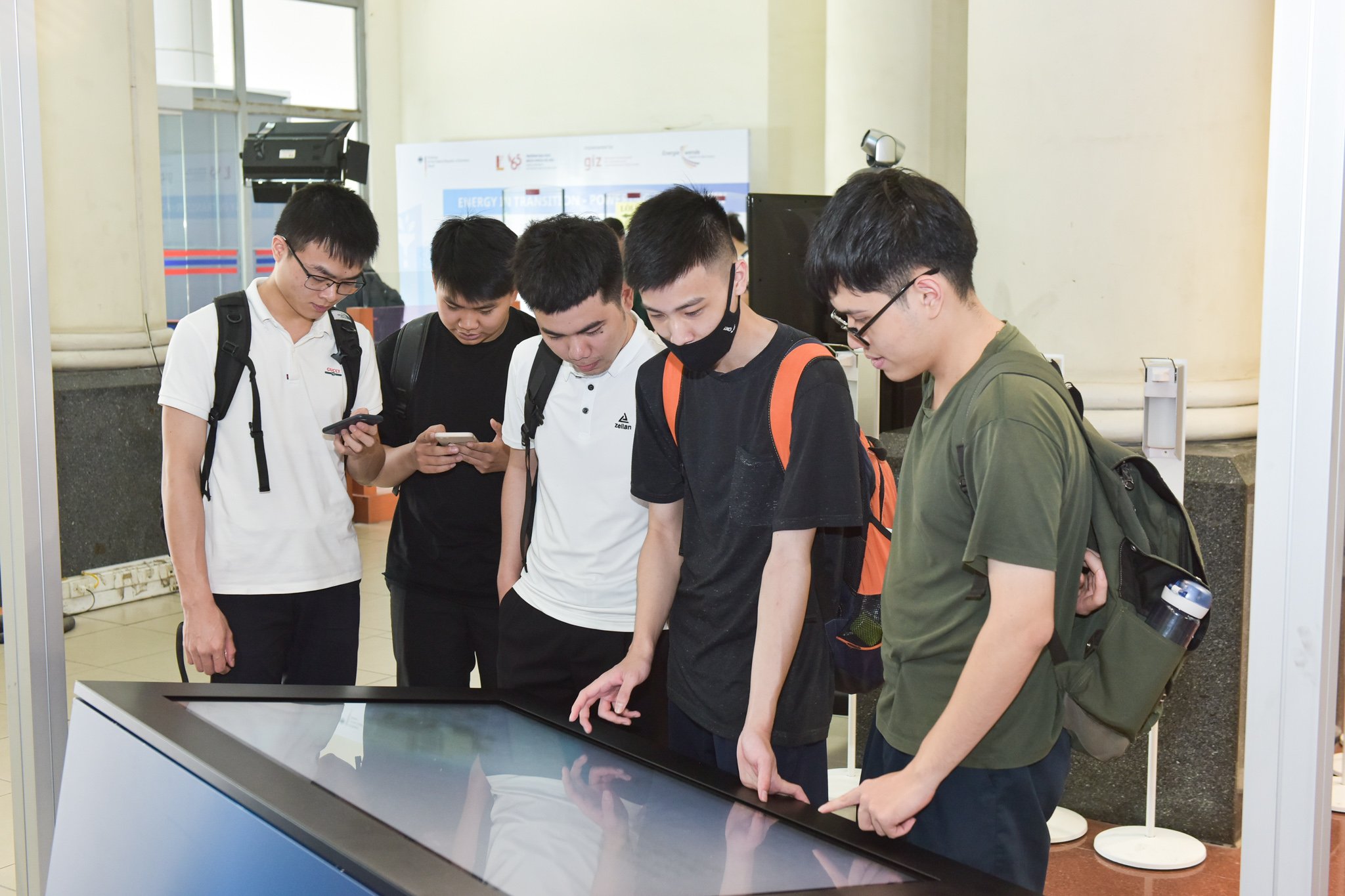 At Hanoi University of Science and Technology (HUST), five male students inform themselves about new mobility concepts at the station "mobility". They are leaning over a waist-high touch screen. April 2021.