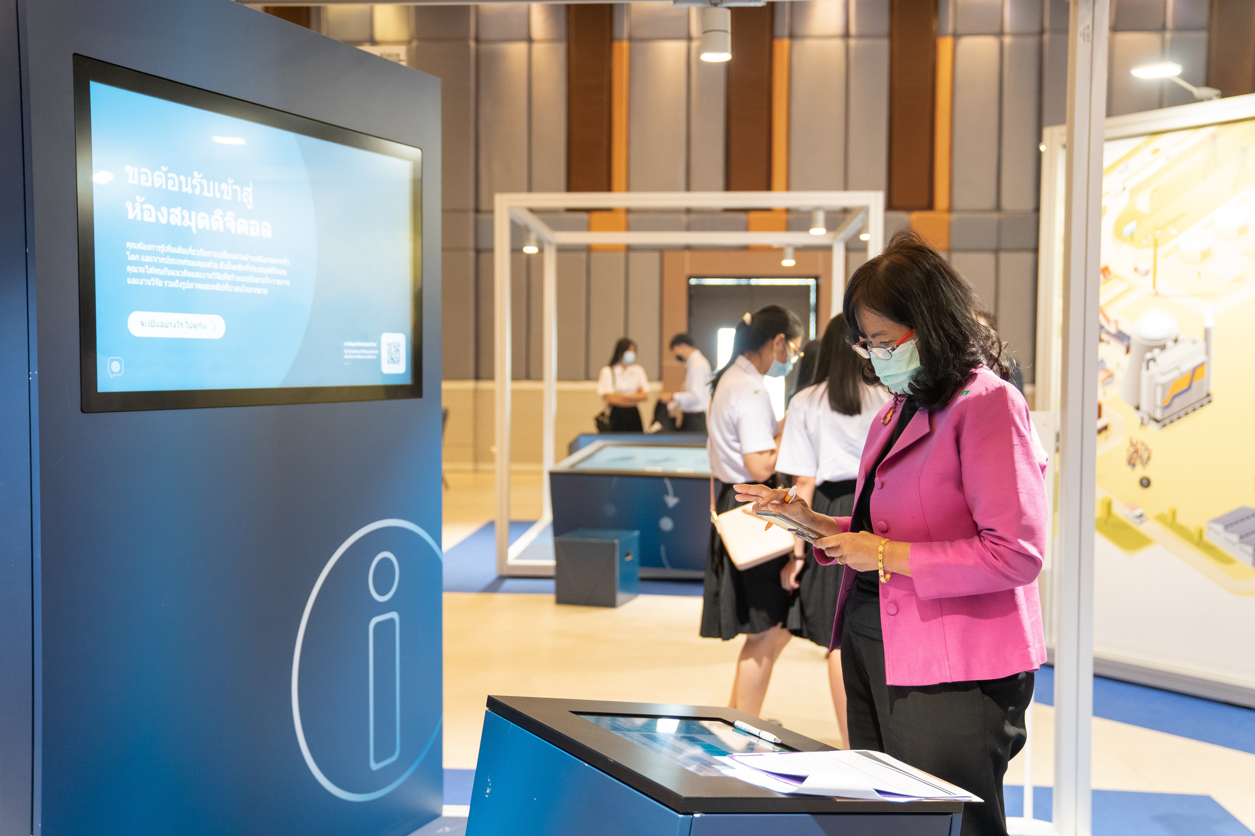 A woman is standing at the "Info Lounge" and looking at her phone. In the background, there are students exploring the station "Mobility" and the "Energy Transition Cube".