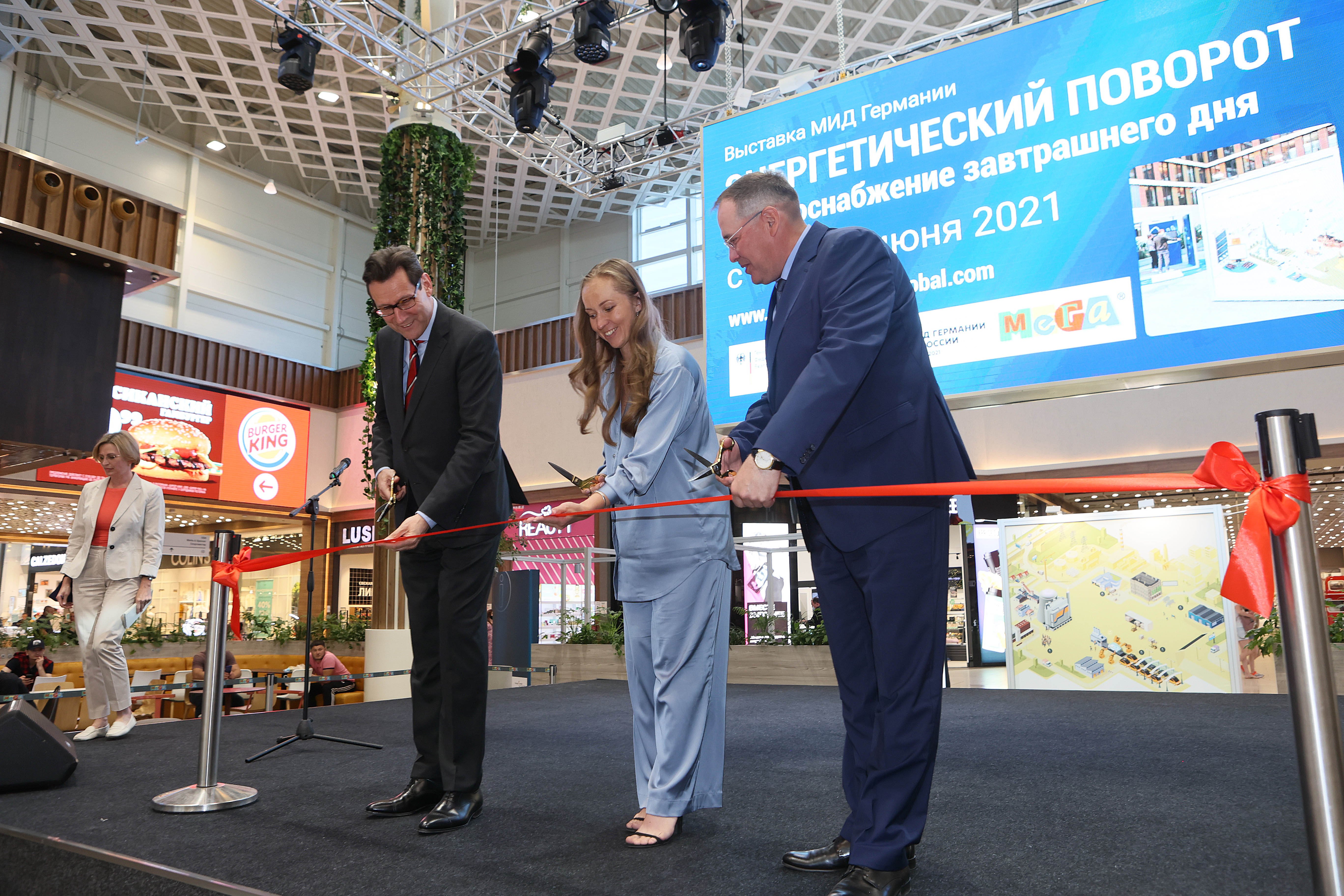 Two men and a woman are cutting a red band to open the travelling exhibition in Yekaterinburg, Russia, while standing on a stage. June 2021.