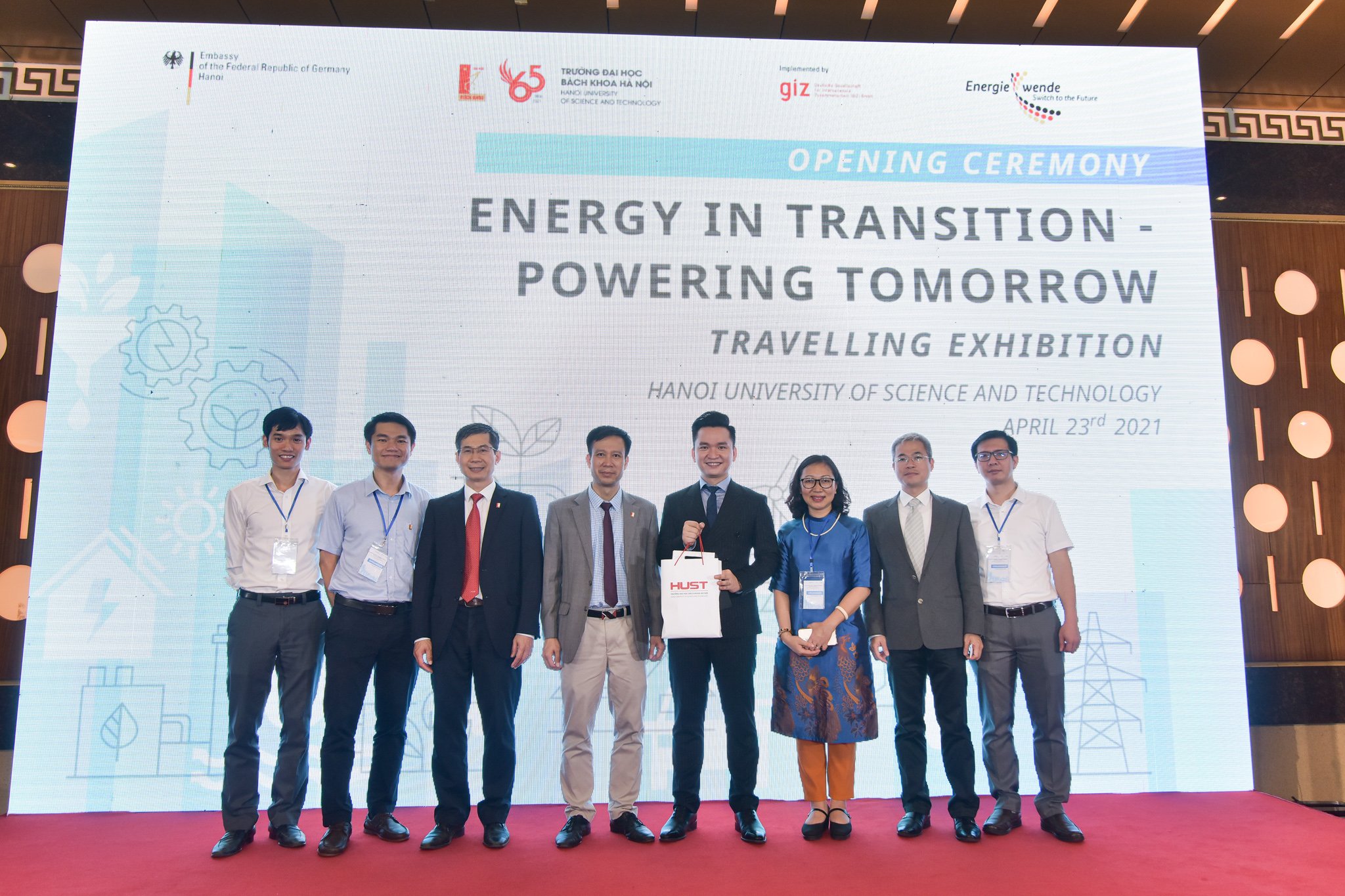 Acht Gäste der Eröffnungszeremonie der Wanderausstellung in Hanoi, Vietnam, stehen auf der Bühne vor einem großen Bildschirm. Auf dem Bildschirm ist der Titel der Ausstellung "Energy in Transition - Powering Tomorrow" geschrieben. April 2021.