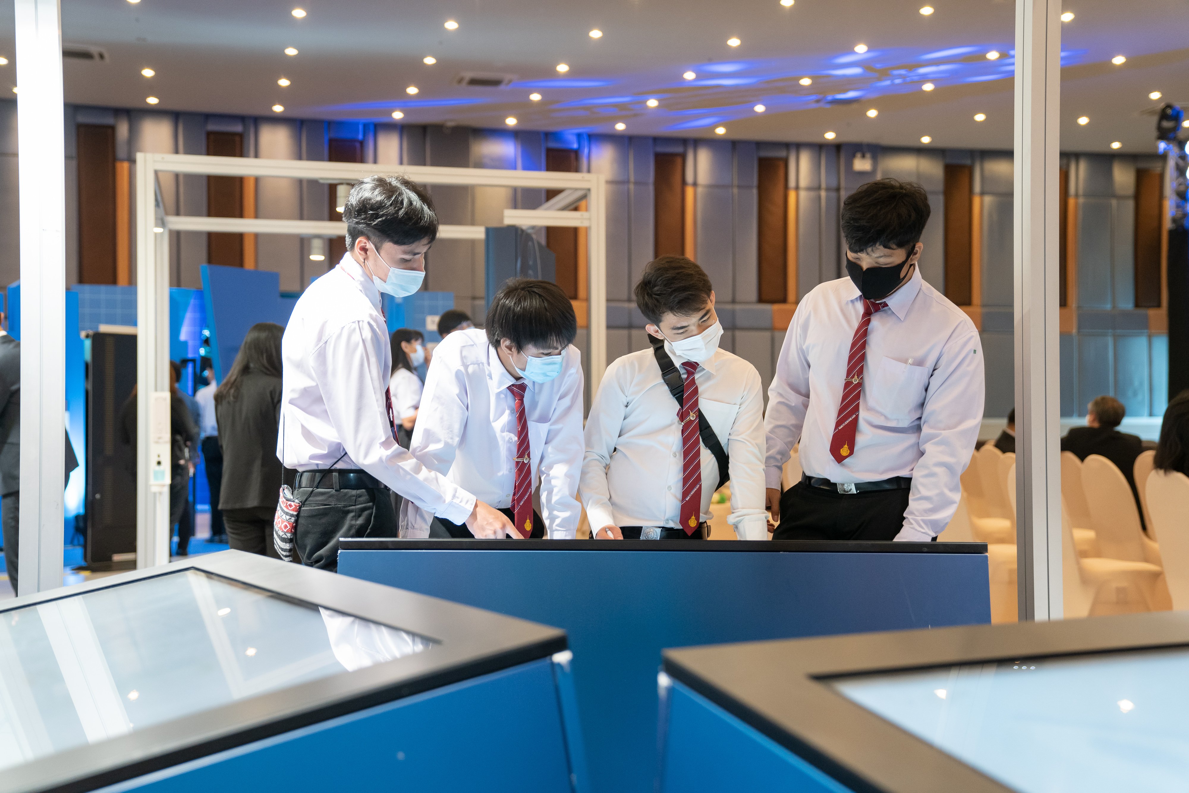 Four male students from Bangkok are standing at blue waist-heigh cube with touch screen of the station "mobility" and inform themselves about the mobility transition. March 2021.