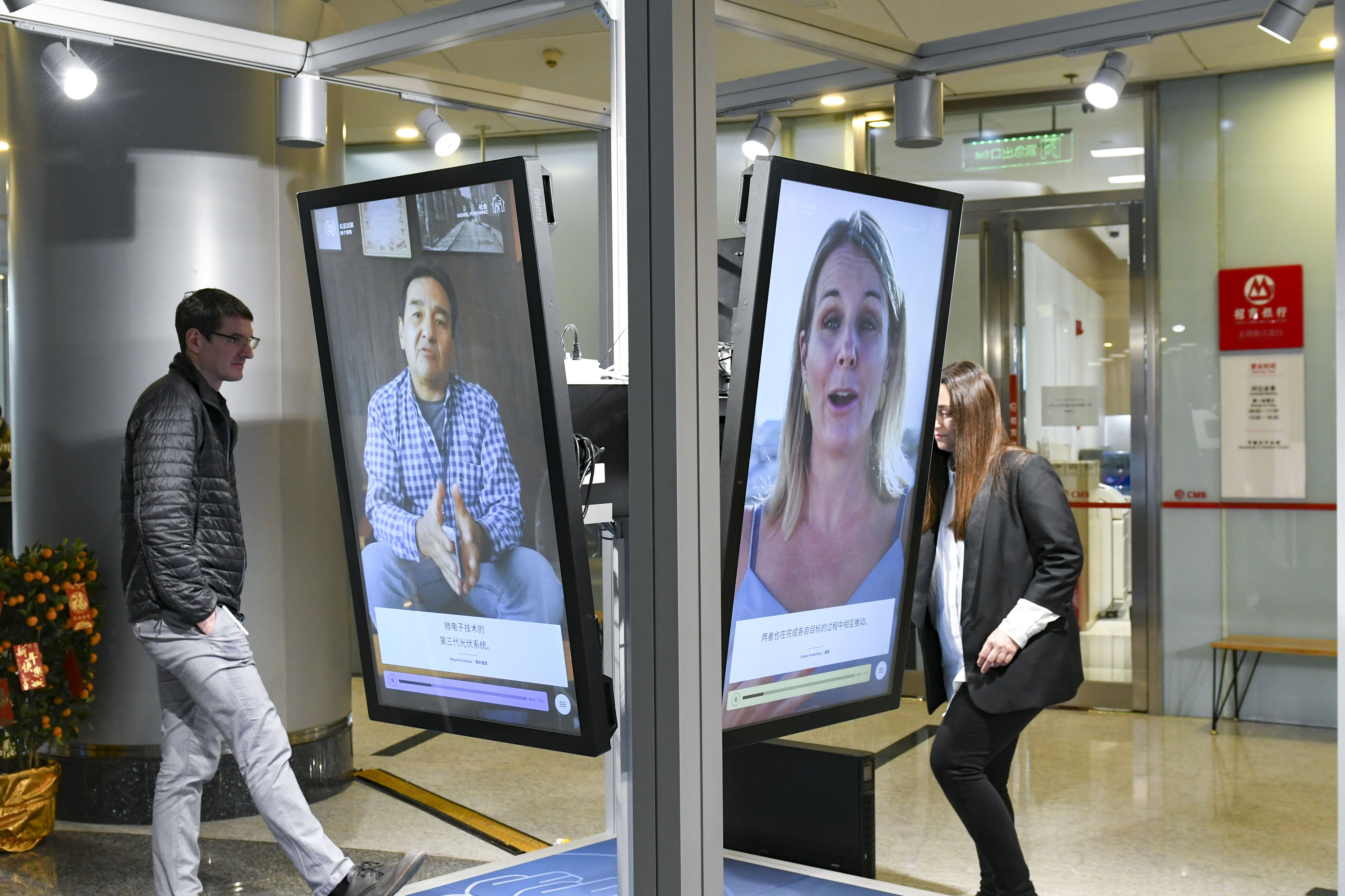 Ein Mann und eine Frau stehen an der Station "Just Transition". Im Vordergrund sind zwei Bildschirme der Station zu sehen. Auf einem Bildschirm spricht ein Mann, auf dem anderen eine Frau. Januar 2021.