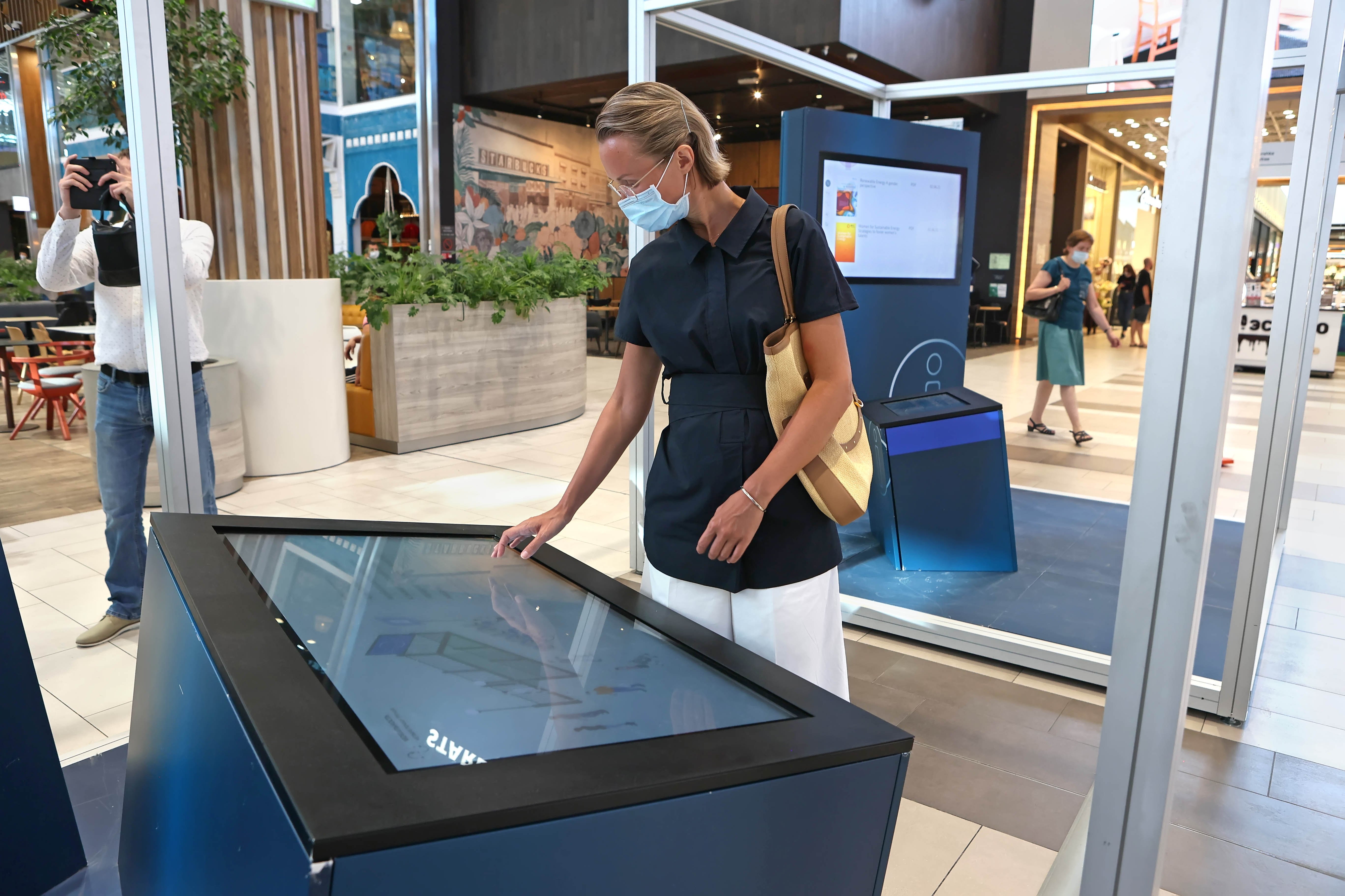 A woman is standing at a waist-high touch screen element of the station "Mobility". She is looking at the screen. In the background, there is the Info Lounge. June 2021.